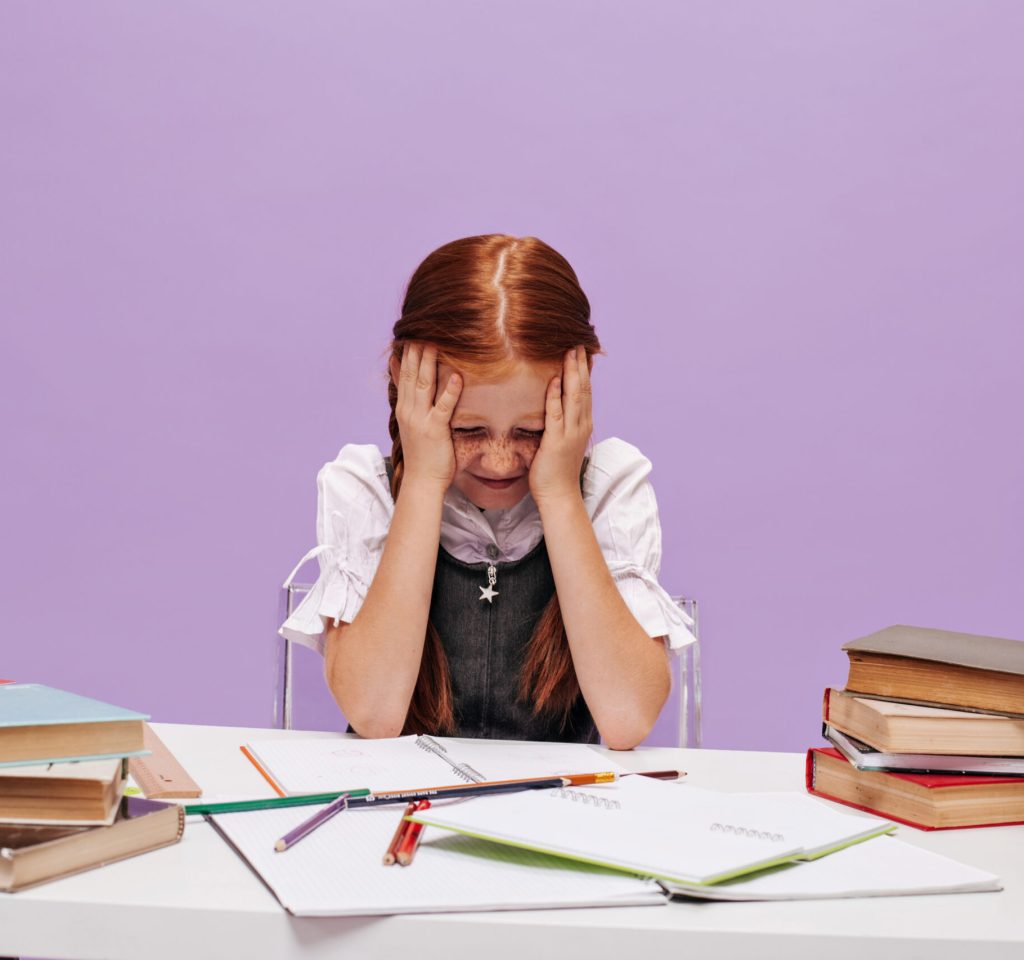 Ginger small girl with freckles in school uniform holding her head with her hands and sitting at desk on purple background..